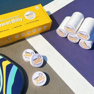 May include: A yellow box with the text "Reusable Compressed Towel Billy" and a white background with a cartoon image of a bear. The box is sitting on a blue and white surface. There are several white compressed towels with the text "Towel Billy" on them.