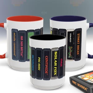 May include: Three white ceramic mugs with black interiors and red, blue, and black handles. Each mug features a different set of vintage video game cartridges printed on the front. The cartridges include titles like "Street Racer", "Solar Fox", "Centipede", "Pac-Man", "Asteroids", "Air Sea Battle", and "Missile Command".