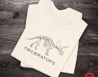 Può includere: Una maglietta bianca con un disegno a linea nera di uno scheletro di Triceratopo. Il testo "TRICERATOPS" è stampato sotto il dinosauro.