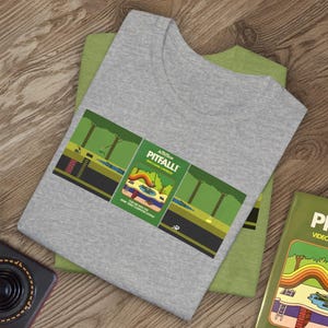 Könnte beinhalten: Ein graues T-Shirt mit einem Vintage-Videospiel-Motiv des Spiels Pitfall! auf der Vorderseite. Das Motiv ist grün, gelb und braun und zeigt eine Dschungelszene mit einer Figur, die ein Seil hält.