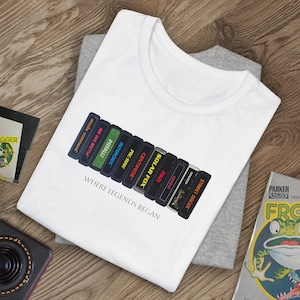 Puede incluir: Camiseta blanca con una pila de cartuchos de videojuegos vintage impresos en la parte delantera. Los cartuchos incluyen títulos como "Street Racer", "Missile Command", "Pitfall" y "Pac-Man". El texto "Where Legends Began" está impreso debajo de los cartuchos.