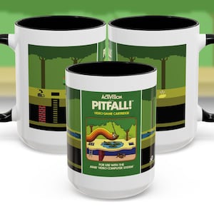 Puede incluir: Una taza de cerámica blanca con un borde y un asa negros. La taza presenta un diseño de cartucho de videojuego retro con el texto "Activision Pitfall! Video Game Cartridge" y una escena de jungla verde. El texto "For use with the Atari Video Computer System" está en la parte inferior.