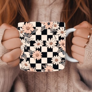 May include: White ceramic mug with a black and cream checkered pattern and daisy floral designs. The daisies have peach-coloured petals and yellow centres. The mug has a white handle and is held by a person.