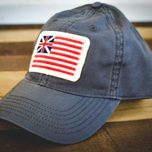 May include: A navy blue baseball cap with a rectangular patch featuring a red and white striped design and a blue and white emblem. The cap has a curved brim and a button on top.