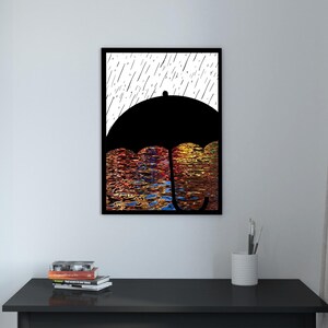 Könnte beinhalten: Gerahmter Kunstdruck mit einer schwarzen Regenschirmsilhouette vor weißem Hintergrund mit Regenlinien. Der untere Teil zeigt farbenfrohe Wasserspiegelungen in Rot, Orange und Gold. Das Kunstwerk wird auf einem schwarzen Tisch ausgestellt.