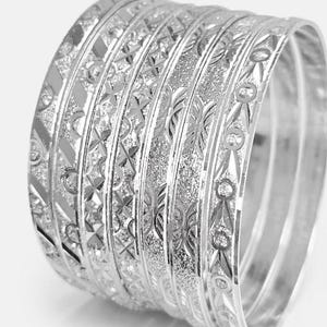 May include: A set of six silver-toned metal bangles featuring a variety of intricate etched patterns, including cross-hatch designs, textured stippled surfaces, and polished geometric shapes that create a reflective and detailed appearance.