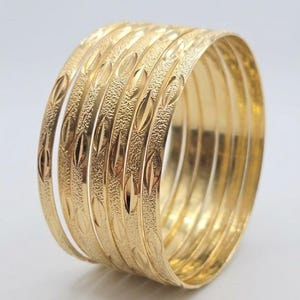 May include: A set of six gold-colored bangles arranged in a stack. Each bangle features a repeating pattern of polished, elongated leaf-shaped motifs set against a textured, matte gold background, creating a consistent metallic appearance.