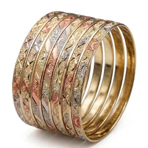 May include: A set of seven stacked bangles featuring a tri-color metal design in yellow, rose, and white tones. Each bangle displays a distinct textured pattern, including cross-hatch, floral, and geometric engravings across the outer surfaces.