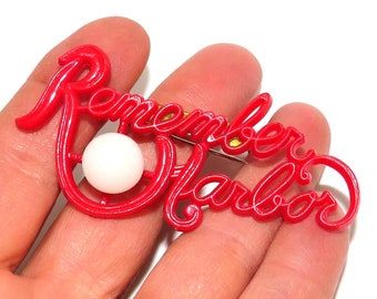 May include: Red plastic brooch with the words "Remember Pearl Harbor" in a stylized script. A white pearl is centered in the brooch.