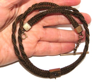 May include: A brown braided necklace with gold-colored metal accents. The necklace is held in a hand, showcasing the intricate braiding and the clasp. The necklace has a T-bar closure and square metal details.
