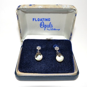 May include: A pair of silver earrings with a white opal stone and a flower design. The earrings are in a blue velvet box with the words "Floating Opals by Milano" printed on the inside.