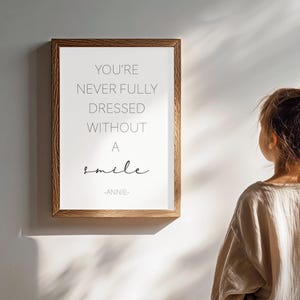 Puede incluir: Un cuadro rectangular con marco de madera de tono medio que presenta el texto YOU'RE NEVER FULLY DRESSED WITHOUT A smile -ANNIE- en letras negras sobre un fondo blanco, exhibido verticalmente en una superficie de pared lisa.