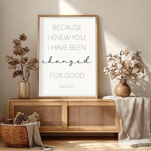 Puede incluir: Una impresión enmarcada con el texto "BECAUSE I KNEW YOU, I HAVE BEEN changed FOR GOOD -WICKED-". La impresión se muestra en un mueble de madera con frente tejido. También hay jarrones decorativos y una cesta con una manta.
