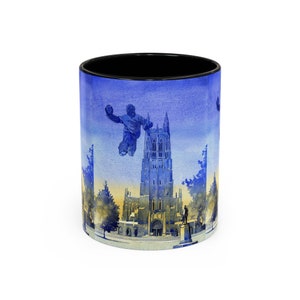 May include: A blue and white ceramic mug featuring a watercolour illustration of a church and a person playing a sport. The church has a tall spire and is surrounded by trees. The person is silhouetted against the sky and is holding a ball.