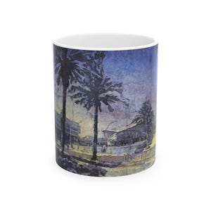 May include: White ceramic coffee mug featuring a watercolor painting of palm trees and buildings under a blue sky. The artwork wraps around the mug, creating a scenic view. The mug is cylindrical and has a white interior.