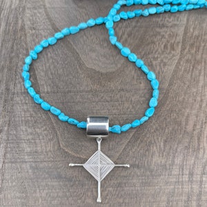 Puede incluir: Un collar de cuentas turquesa con un colgante de plata con un diseño de cruz envuelto en alambre.