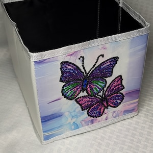 May include: A white fabric storage box with a silver trim. The box has a colorful butterfly design with purple, pink, green, and blue accents.