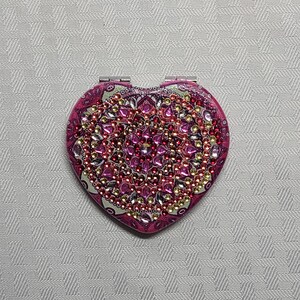 Pode incluir: Um espelho de bolso em forma de coração com um design de mandala rosa e verde. O mandala é decorado com strass rosa, vermelho, dourado e transparente.