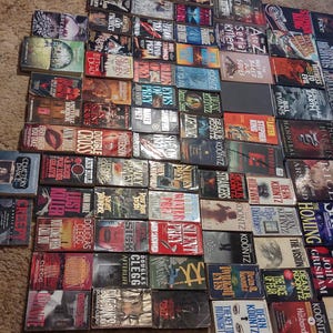 K&ouml;nnte beinhalten: Eine gro&szlig;e Sammlung von Taschenb&uuml;chern, die in einem Rastermuster auf einer strukturierten Oberfl&auml;che angeordnet sind. Die B&uuml;cher zeigen verschiedene Cover-Designs, Titel und Autorennamen, darunter Werke von Dean Koontz, John Saul und John Grisham.