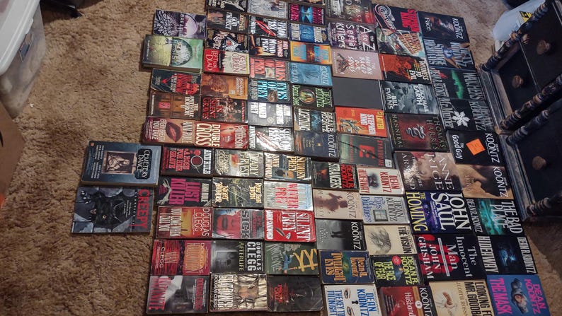 K&ouml;nnte beinhalten: Eine gro&szlig;e Sammlung von Taschenb&uuml;chern, die in einem Rastermuster auf einer strukturierten Oberfl&auml;che angeordnet sind. Die B&uuml;cher zeigen verschiedene Cover-Designs, Titel und Autorennamen, darunter Werke von Dean Koontz, John Saul und John Grisham.