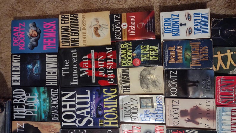K&ouml;nnte beinhalten: Eine Sammlung von Taschenb&uuml;chern, die in einem Raster angeordnet sind, mit Titeln von Dean Koontz, John Grisham und John Saul. Sichtbare Titel sind unter anderem The Mask, Looking for Mr. Goodbar, The Husband, The Key to Midnight, Hideaway und The Homing.