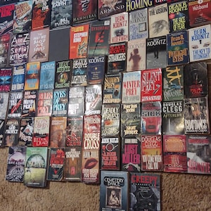 K&ouml;nnte beinhalten: Eine gro&szlig;e Sammlung von Taschenb&uuml;chern aus den Bereichen Thriller und Horror, die in einem Raster an einer Wand angeordnet sind. Die B&uuml;cher zeigen verschiedene Titel und Autoren wie Dean Koontz, John Sandford und Douglas Clegg mit bunten Covern.