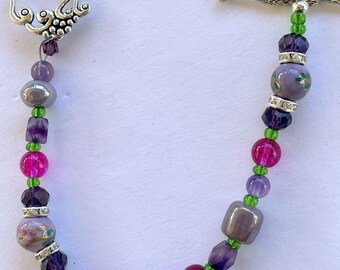Puede incluir: Un collar de cuentas moradas, rosas y verdes con un cierre plateado. El collar presenta una variedad de cuentas, incluyendo formas redondas, cuadradas y ovaladas. Las cuentas están intercaladas con pequeñas cuentas plateadas.