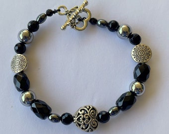 Puede incluir: Un brazalete negro y plateado con un dije en forma de corazón y cuentas con patrones geométricos.