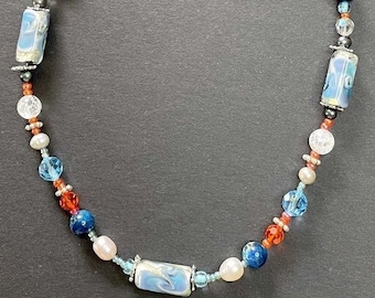 Puede incluir: Un collar de cuentas multicolores con un cierre plateado. El collar presenta cuentas azules, blancas, naranjas y rosas, así como cuentas plateadas y negras. El collar está hecho de varios materiales, incluyendo vidrio, metal y perlas.
