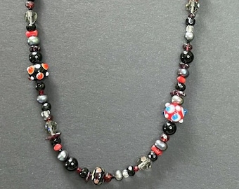 Puede incluir: Un collar de cuentas negras y rojas con un cierre plateado. El collar presenta varias cuentas, incluyendo cuentas negras, rojas y plateadas, así como algunas con un diseño estampado.