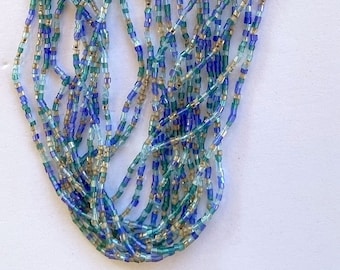 Puede incluir: Un collar de múltiples hilos hecho de pequeñas cuentas cuadradas en tonos azules, verdes y beige. El collar tiene un cierre plateado.