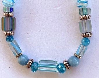 Puede incluir: Un brazalete de cuentas azules y plateadas con cierre de palanca. Las cuentas son una mezcla de vidrio transparente, azul y blanco, y algunas tienen un patrón a rayas.