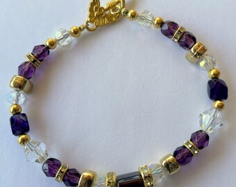 Puede incluir: Un brazalete de oro con cuentas de vidrio morado y transparente. El brazalete tiene un cierre de oro con un diseño floral.