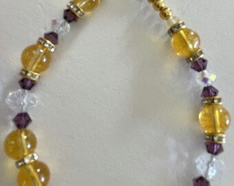 Puede incluir: Un brazalete de tono dorado con cierre de palanca. El brazalete está hecho con cuentas facetadas amarillas y transparentes y cuentas moradas.