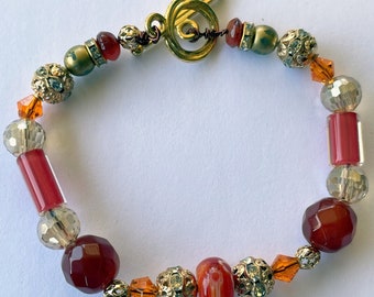 Puede incluir: Un brazalete de tono dorado con cierre de palanca presenta cuentas de vidrio rojas, naranjas y transparentes. El brazalete tiene una mezcla de cuentas redondas, facetadas y rectangulares.