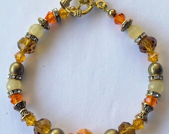 Puede incluir: Un brazalete de tono dorado con una variedad de cuentas de vidrio ámbar, naranja y amarillo. El brazalete tiene un cierre de tono dorado y una sola cuenta redonda con un patrón floral.