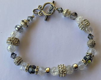 Puede incluir: Un brazalete plateado con cuentas de cristal transparente y gris, un cierre de palanca plateado y una cuenta plateada con pedrería.
