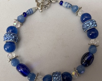 Puede incluir: Una pulsera de cuentas azules con una mezcla de cuentas redondas, facetadas y ovaladas. La pulsera incluye elementos de cristal y dos cuentas espaciadoras con incrustaciones de pedrería. Se cierra con un cierre de palanca plateado.