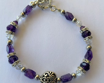 Puede incluir: Un brazalete de plata con un dije en forma de corazón y cuentas de amatista púrpura. El brazalete también presenta cuentas de cristal transparente y separadores de plata.