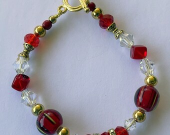 Puede incluir: Una pulsera de cuentas rojas, doradas y transparentes, con un cierre de palanca dorado. Las cuentas incluyen cubos rojos facetados, cristales transparentes y cuentas metálicas doradas. Algunas cuentas rojas tienen rayas doradas.