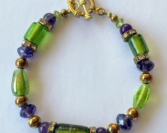 Puede incluir: Un brazalete de cuentas verdes y moradas con un cierre dorado. El brazalete presenta cuentas de vidrio verdes y moradas, cuentas doradas y pedrería transparente.