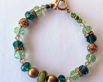 Puede incluir: Un brazalete de cuentas verdes y doradas con un cierre de palanca dorado. El brazalete presenta cuentas de vidrio verdes y transparentes, cuentas doradas y algunas cuentas doradas más grandes con una superficie texturizada.