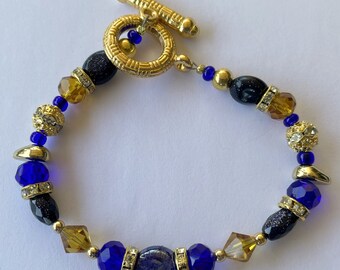 Puede incluir: Un brazalete de cuentas doradas y azules con un cierre de palanca dorado. El brazalete presenta cuentas azules y doradas, así como cuentas negras con un acabado brillante.
