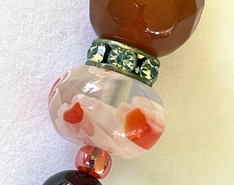 Puede incluir: Primer plano de un collar de cuentas multicolores. Las cuentas están hechas de cornalina, ágata y vidrio. Las cuentas de cornalina son de color naranja intenso, las cuentas de ágata son de color marrón claro y las cuentas de vidrio son de color blanco con remolinos rojos y naranjas. Las cuentas están separadas por un separador de metal plateado con diamantes de imitación.