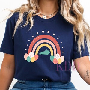 May include: A navy blue t-shirt with a rainbow design featuring a stylized outline of the state of Kentucky. The rainbow is made up of red, pink, yellow, and blue stripes. The rainbow is surrounded by white dots and has colorful Easter eggs on either side.