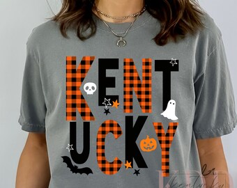 May include: A grey t-shirt with the word "KENTUCKY" in black letters with orange and black buffalo check pattern. The letters are decorated with Halloween-themed graphics, including a skull, a ghost, a bat, and a jack-o-lantern.