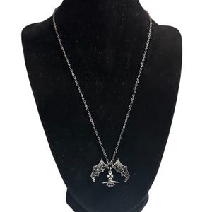 May include: A silver-toned necklace featuring a pendant shaped like a planet with a cross on top, flanked by two bat wings. The wings are embellished with small, dark, sparkling stones. The pendant hangs from a delicate silver-toned metal chain.