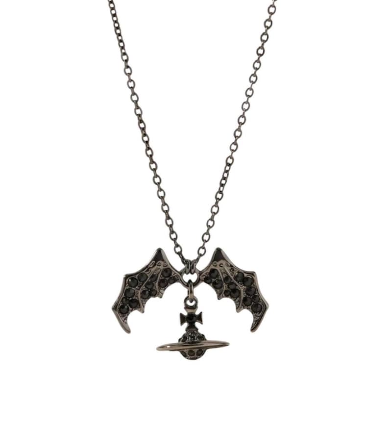 May include: A dark gunmetal-toned necklace featuring a pendant shaped like a planet with a cross on top, flanked by two bat-like wings. The wings and the central orb are encrusted with small, dark, reflective crystals, suspended from a thin chain.
