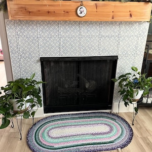 May include: An oval braided rug in shades of purple, green, pink, and white sits in front of a fireplace. The fireplace has a black screen and is surrounded by plants in white pots on black stands. A wooden mantel is decorated with greenery and gnomes.