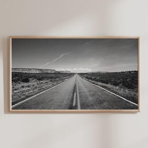 Puede incluir: Fotografía en blanco y negro enmarcada de una carretera larga y recta que se extiende en la distancia bajo un cielo nublado. La carretera está bordeada de vegetación baja y colinas distantes.